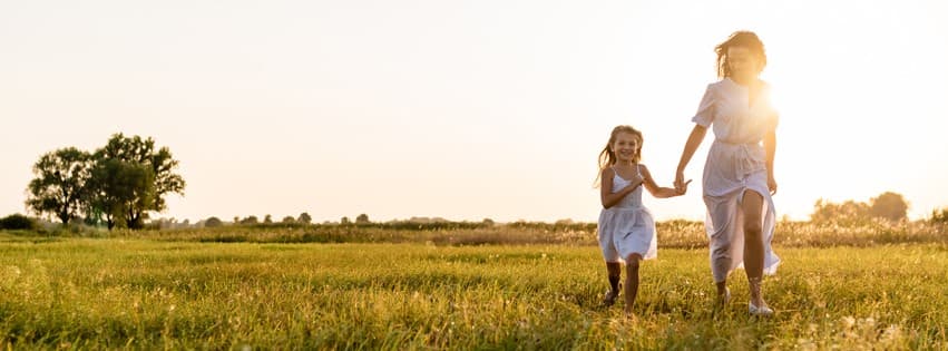 A woman and a young girl, both wearing white dresses, hold hands and run through a sunlit grassy field. The sun is low in the sky, casting a warm, golden glow over the scene.
