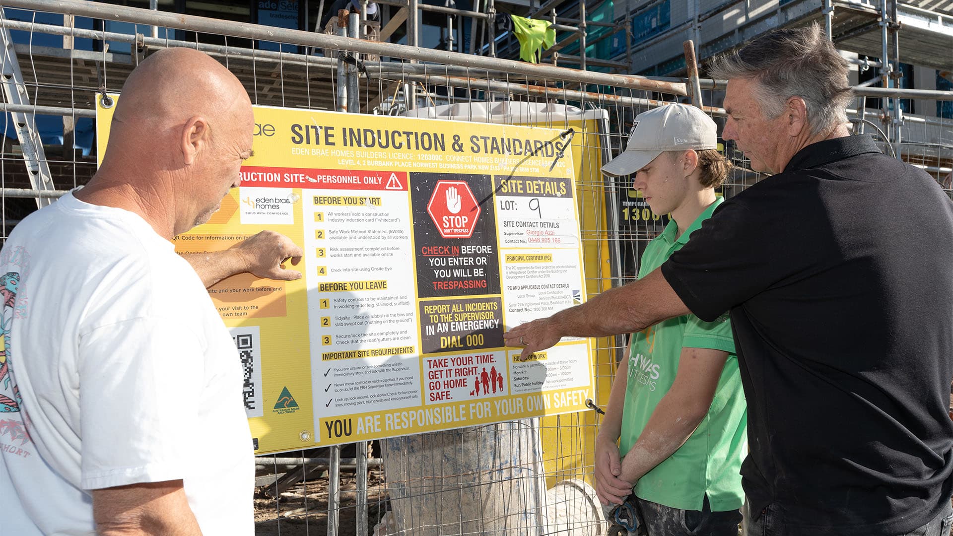 Three men stand in front of a construction site sign, with one man pointing to information on the board while the others look on attentively, surrounded by scaffolding and safety gear on a sunny day.