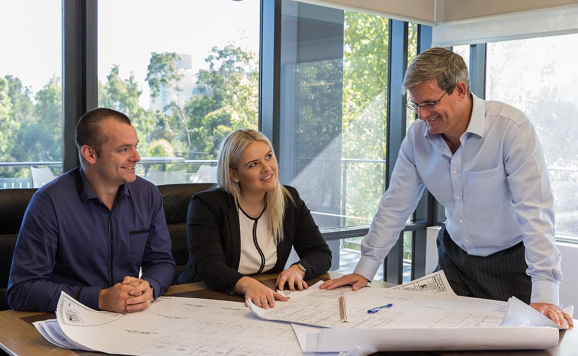 Three people sit and stand around a table covered with architectural blueprints, engaged in discussion. Large windows behind them reveal trees and natural light fills the modern office space.