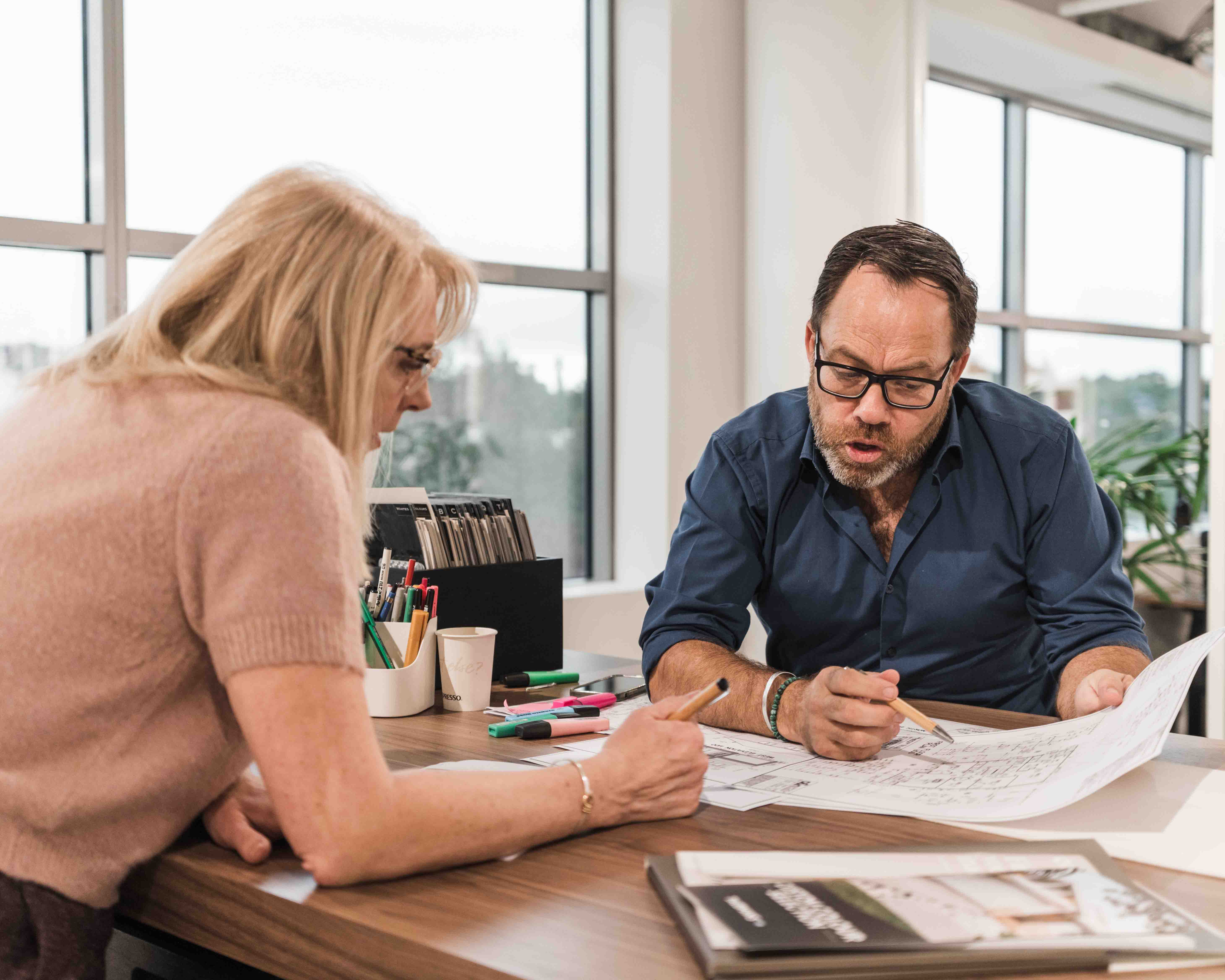 Two people sit at a desk in a modern office, reviewing architectural plans. The man, wearing glasses and a blue shirt, gestures to the plans, while the woman, with blonde hair and a pink top, makes notes.