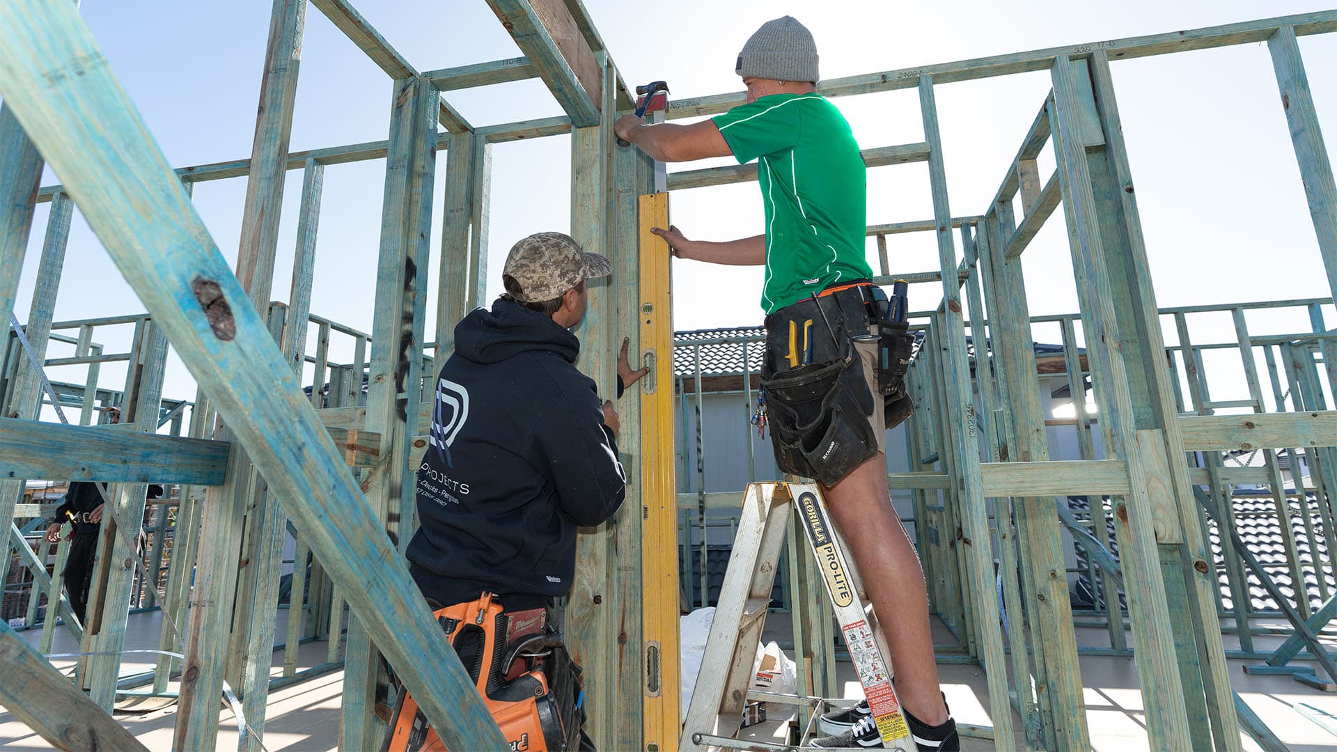 Two construction workers build a wooden house frame; one stands on a ladder using a power drill, while the other holds a spirit level to align the vertical beam. Sunlight streams through the unfinished structure.