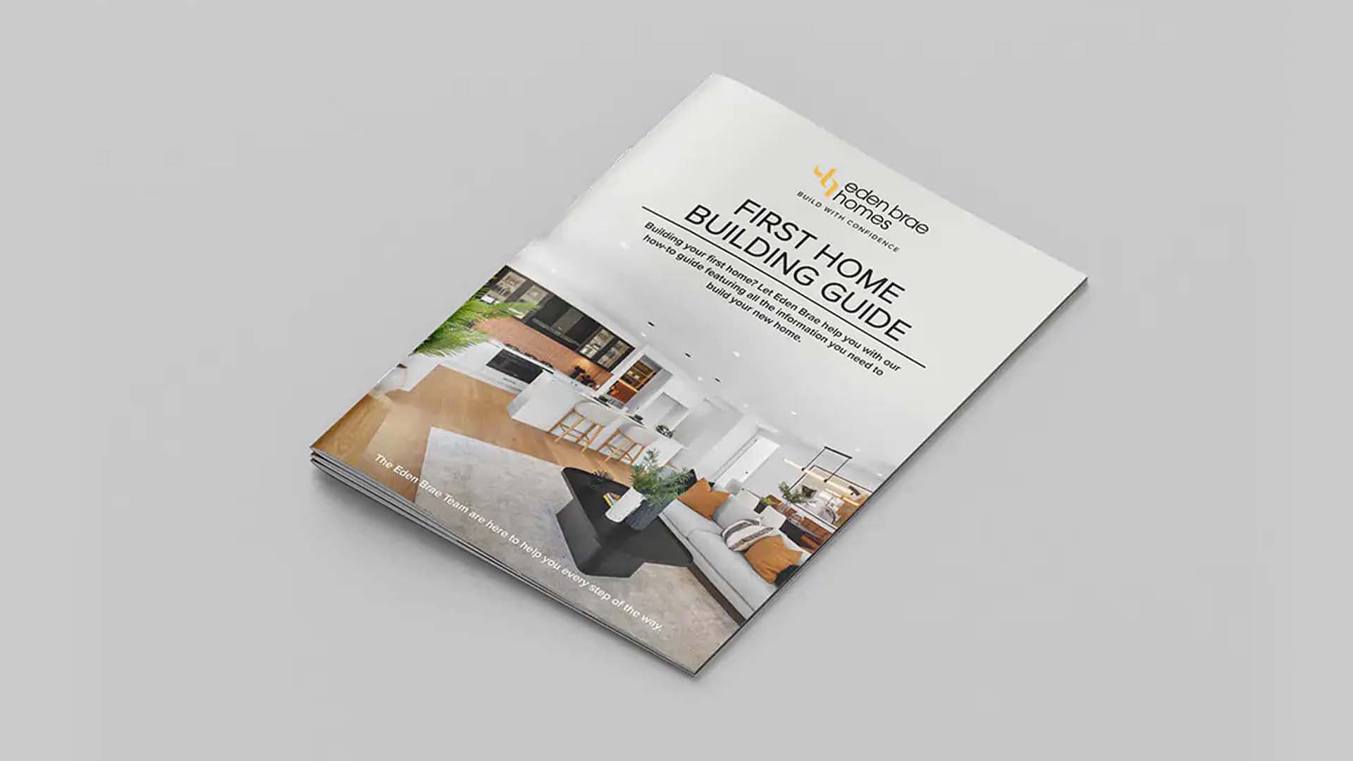 A brochure titled First Home Building Guide with a modern living room image on the cover, featuring a white kitchen, sofa, and green plant, placed on a plain, light gray surface.