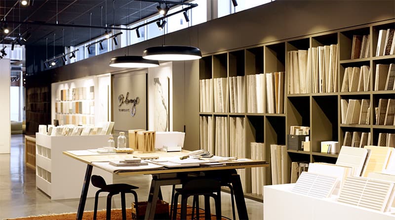 A modern showroom with shelves filled with tile samples, a large table with chairs, overhead pendant lights, and neatly arranged design materials creating a stylish and organized workspace.