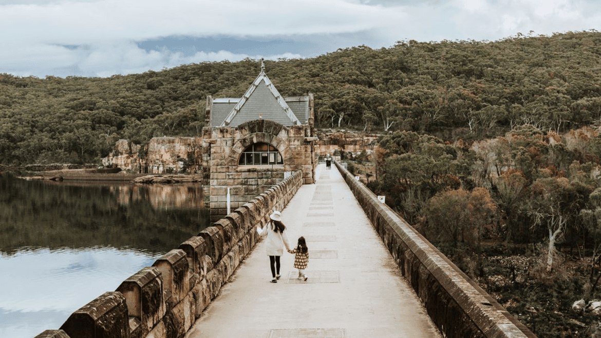 A woman and a young girl walk hand in hand along a stone bridge over water, heading toward a historic stone building, with dense green forest and hills in the background.