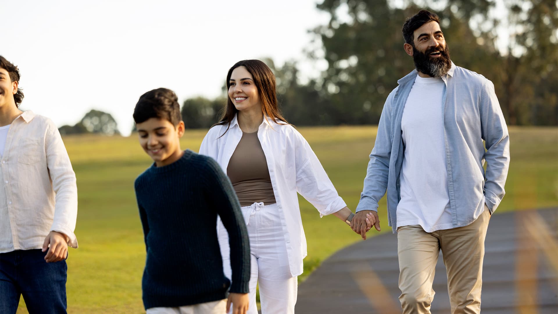 A smiling family of four, including two adults and two children, walks hand in hand outdoors on a sunny day, with green grass and trees in the background.