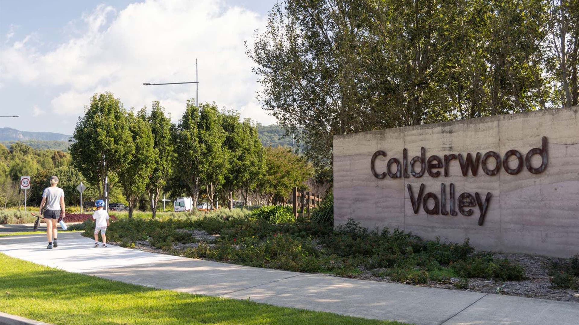 A woman and a child walk on a path beside a large concrete sign that reads Calderwood Valley, surrounded by greenery and trees under a partly cloudy sky.