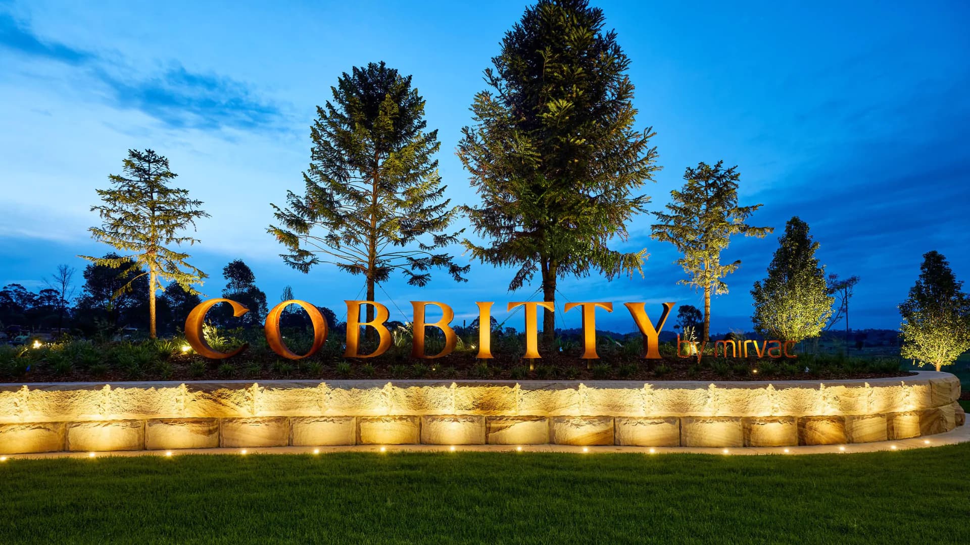 Illuminated sign reading COBBITTY with smaller text below, set against landscaped greenery and tall trees at dusk, with spotlights highlighting the stone and metal structure.