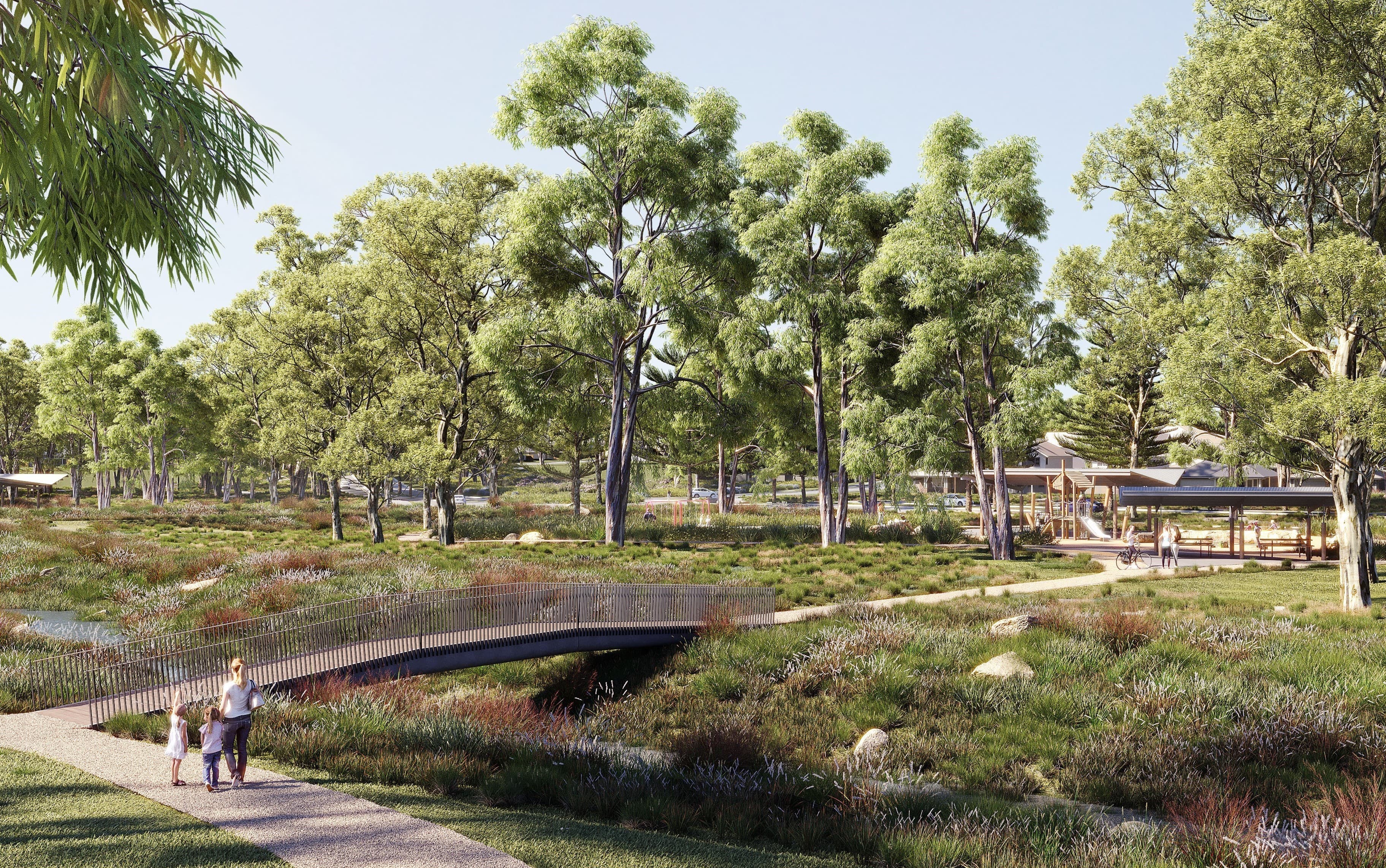 A family walks along a curved path toward a small footbridge in a park filled with tall trees, grassy plants, and a picnic shelter in the background under a clear blue sky.