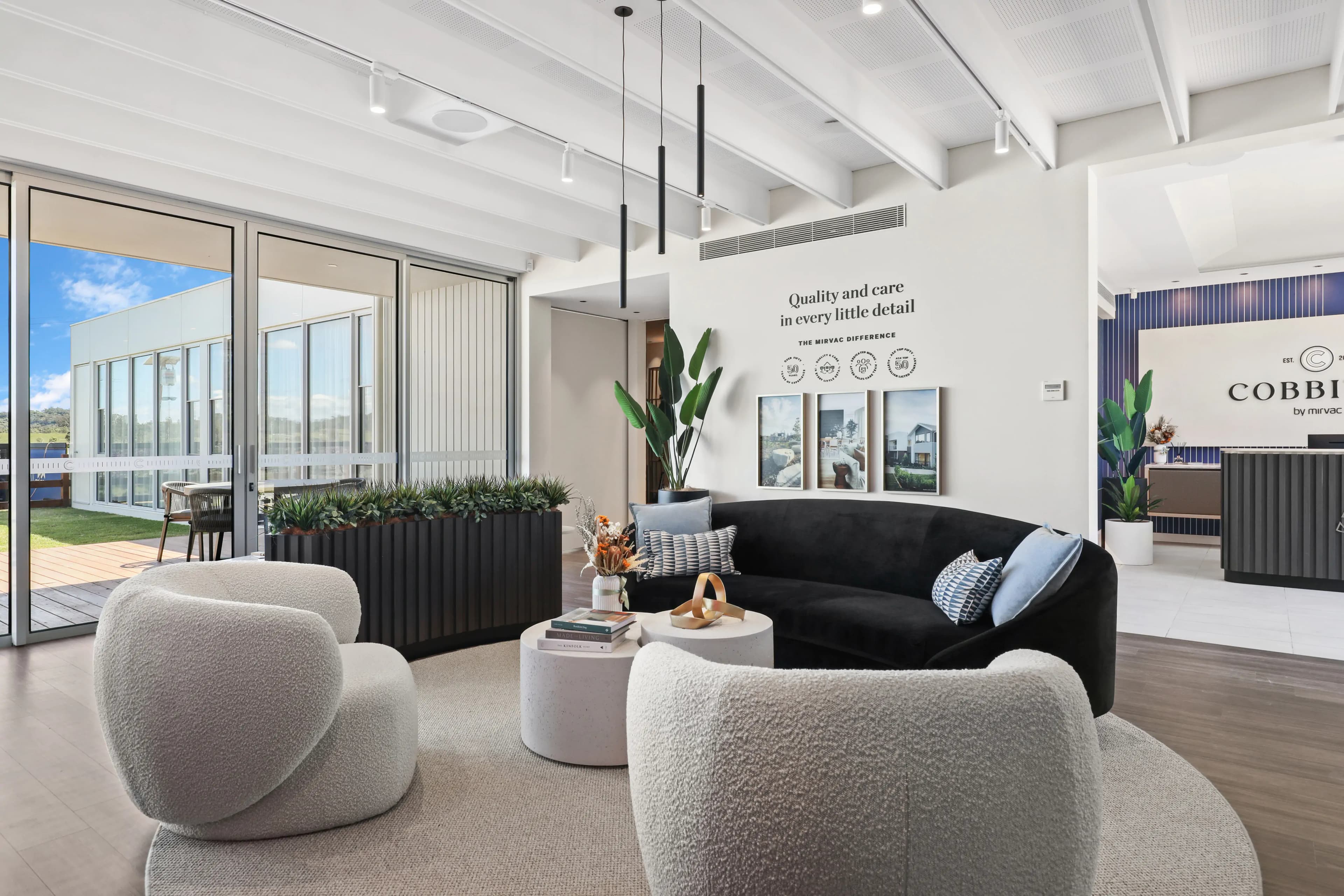 Modern office lounge with black velvet sofa, round white chairs, coffee tables, indoor plants, and large windows letting in natural light. Wall art displays the phrase “Quality and care in every little detail.”.