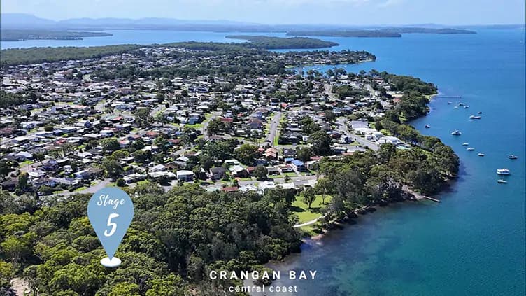 Aerial view of a coastal suburb with houses surrounded by greenery, a shoreline with boats anchored in the water, and a marker labeled Stage 5 over Crangan Bay, Central Coast.