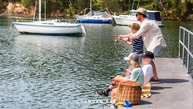 A man and three children sit and stand on a dock, fishing by the water. Sailboats float in the background, and a picnic basket is beside them. The scene is bright and peaceful, suggesting a family outing.