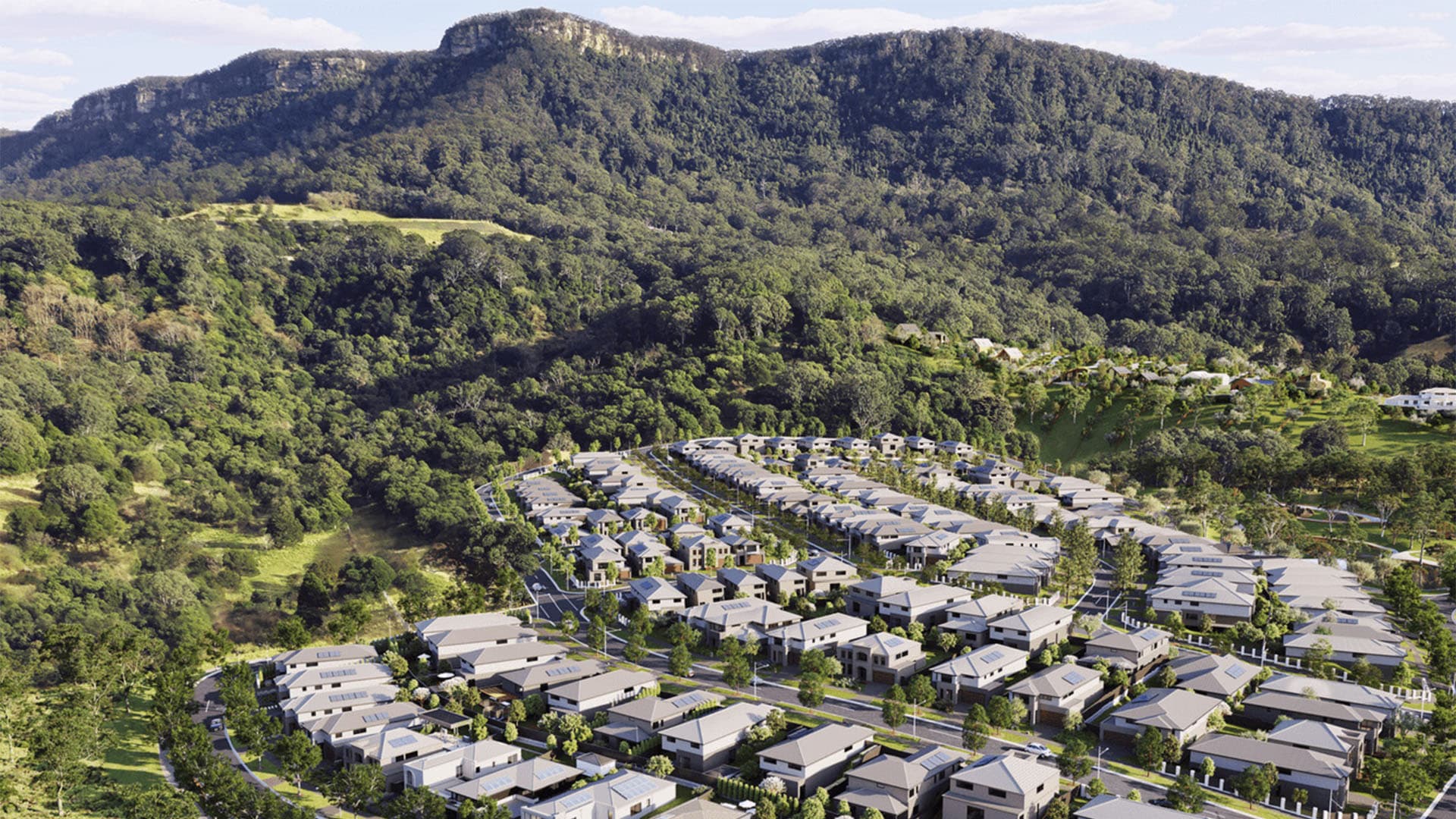 Aerial view of a suburban neighborhood with rows of modern houses surrounded by dense green forest and hills, with a tall, tree-covered mountain in the background under a partly cloudy sky.