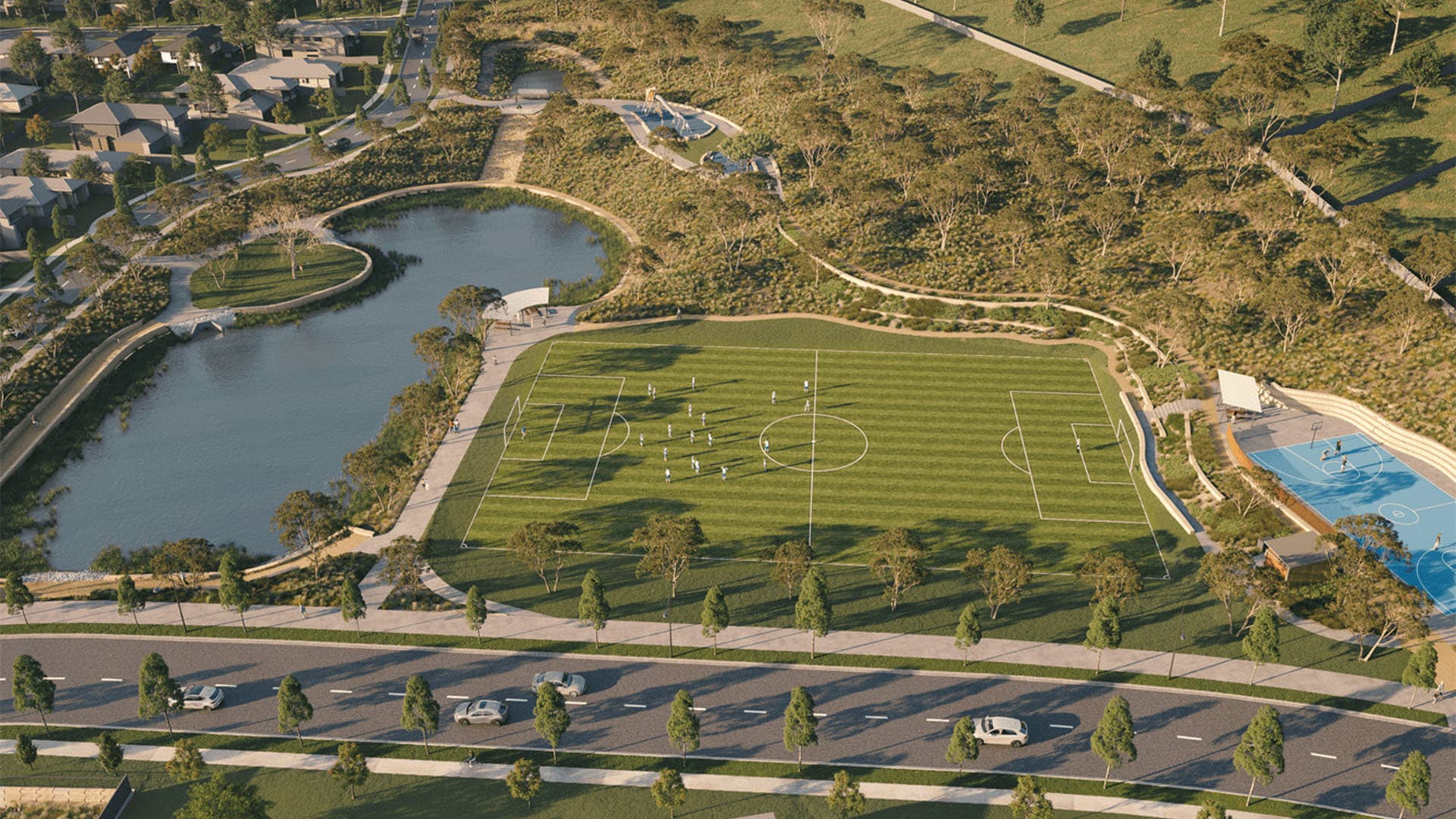 Aerial view of a green soccer field with players, next to a blue basketball court, a pond, walking paths, and nearby houses surrounded by trees and greenery. Cars are parked along the road in the foreground.