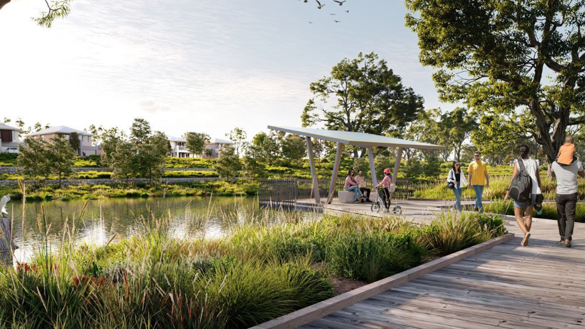 People walk along a wooden boardwalk beside a pond surrounded by greenery. Some stop at a modern shelter, while others stroll with children. Houses are visible in the background under a bright sky.