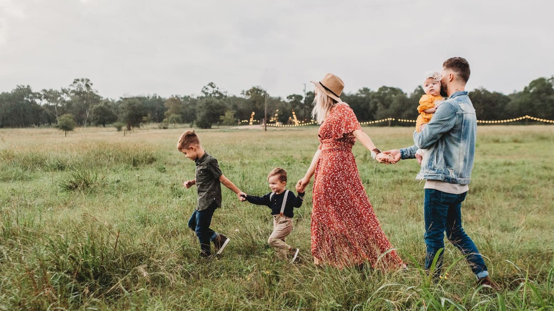 A family of five walks hand in hand through a grassy field. The mother wears a red dress and hat, the father a denim jacket, and they are with three young children. Trees and string lights are visible in the background.