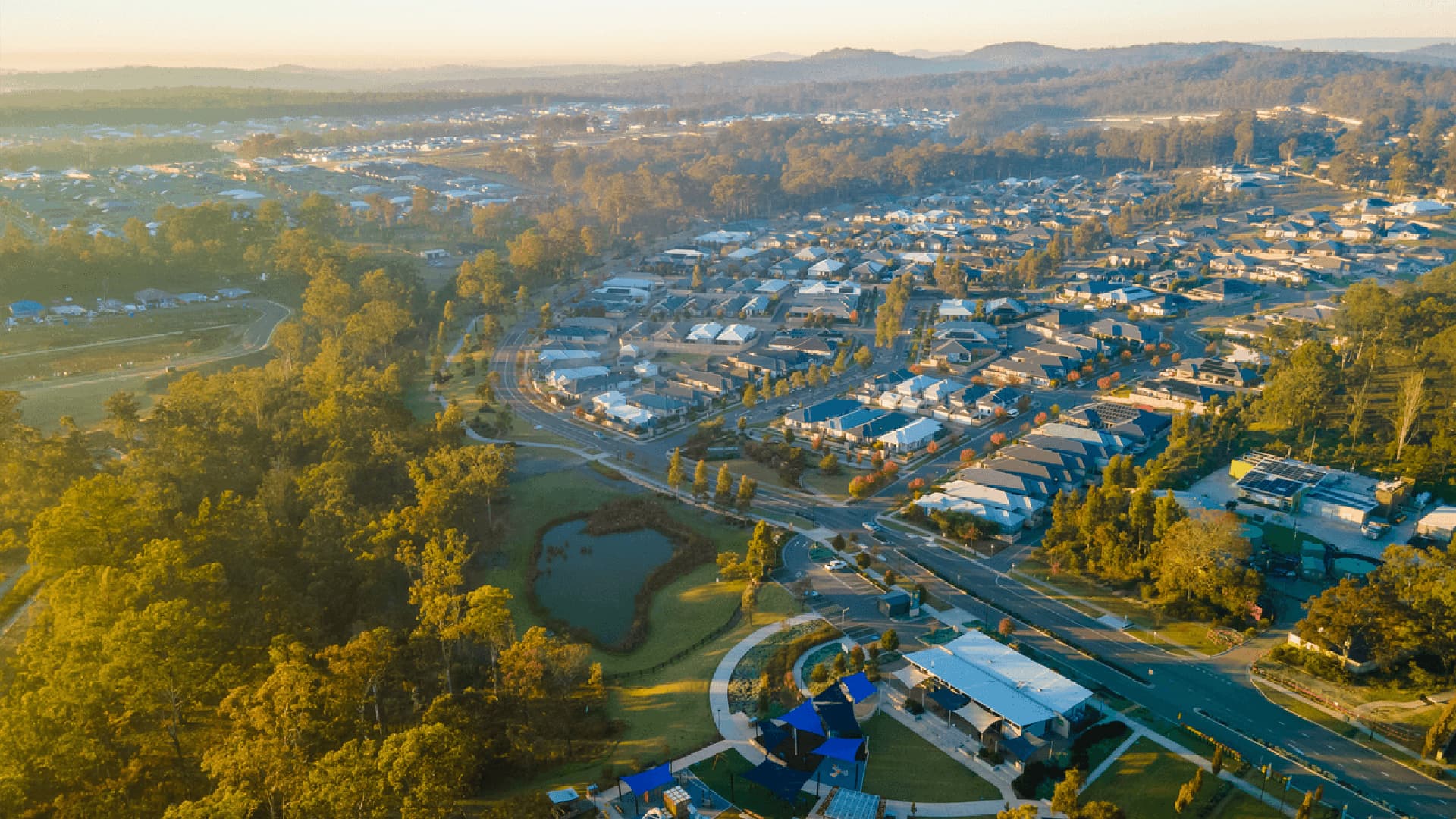 Aerial view of a suburban neighborhood with rows of houses, winding roads, green parks, trees, and a small pond, set against a backdrop of hills under a hazy sky at sunrise.