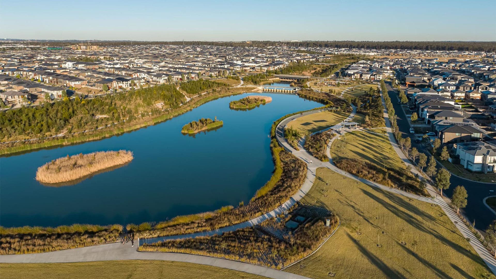 Aerial view of a suburban neighborhood with houses surrounding a large, winding lake featuring small islands and walking paths, bordered by green spaces and open fields under a clear blue sky.