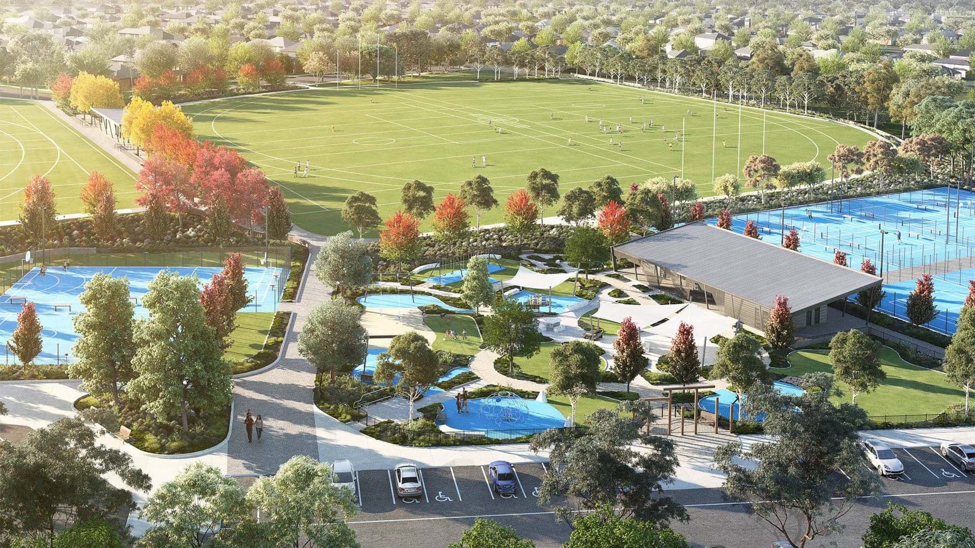 Aerial view of a large sports complex with green fields, tennis courts, parking area, trees with autumn colors, pathways, and a modern building surrounded by landscaped gardens. People are playing sports and walking.