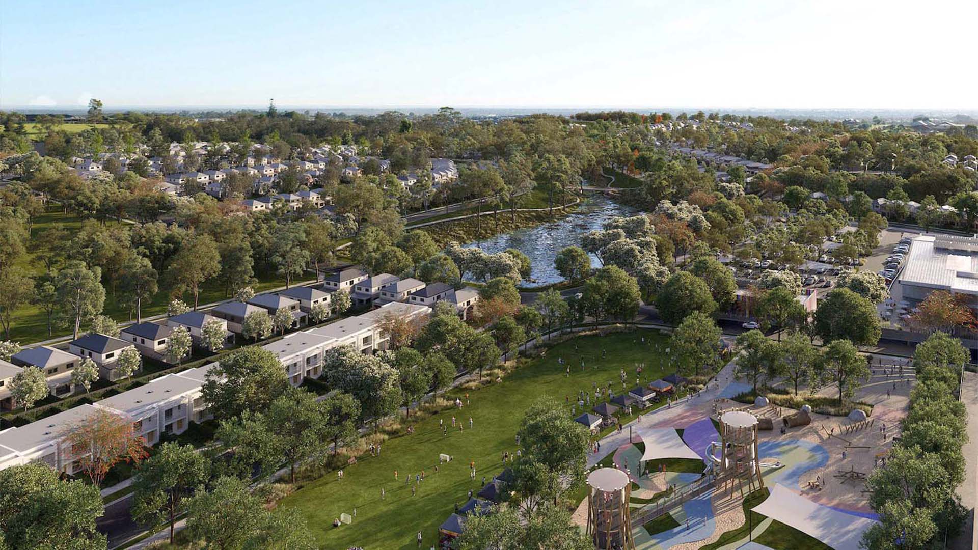 Aerial view of a master-planned community with modern houses, green parks, a pond, walking paths, playgrounds, and a parking lot, surrounded by dense trees and open grassy areas under a clear sky.