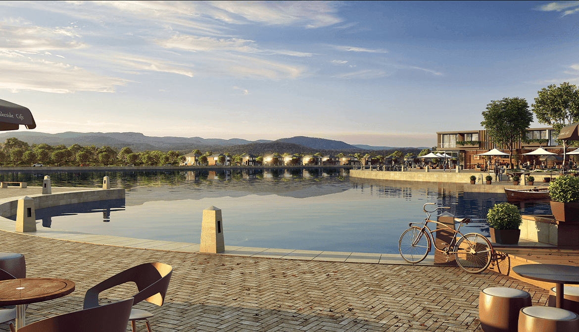 A serene waterfront scene with calm water, outdoor seating, potted plants, and a parked bicycle in the foreground. Buildings, umbrellas, and trees surround the water, with mountains and a partly cloudy sky in the background.