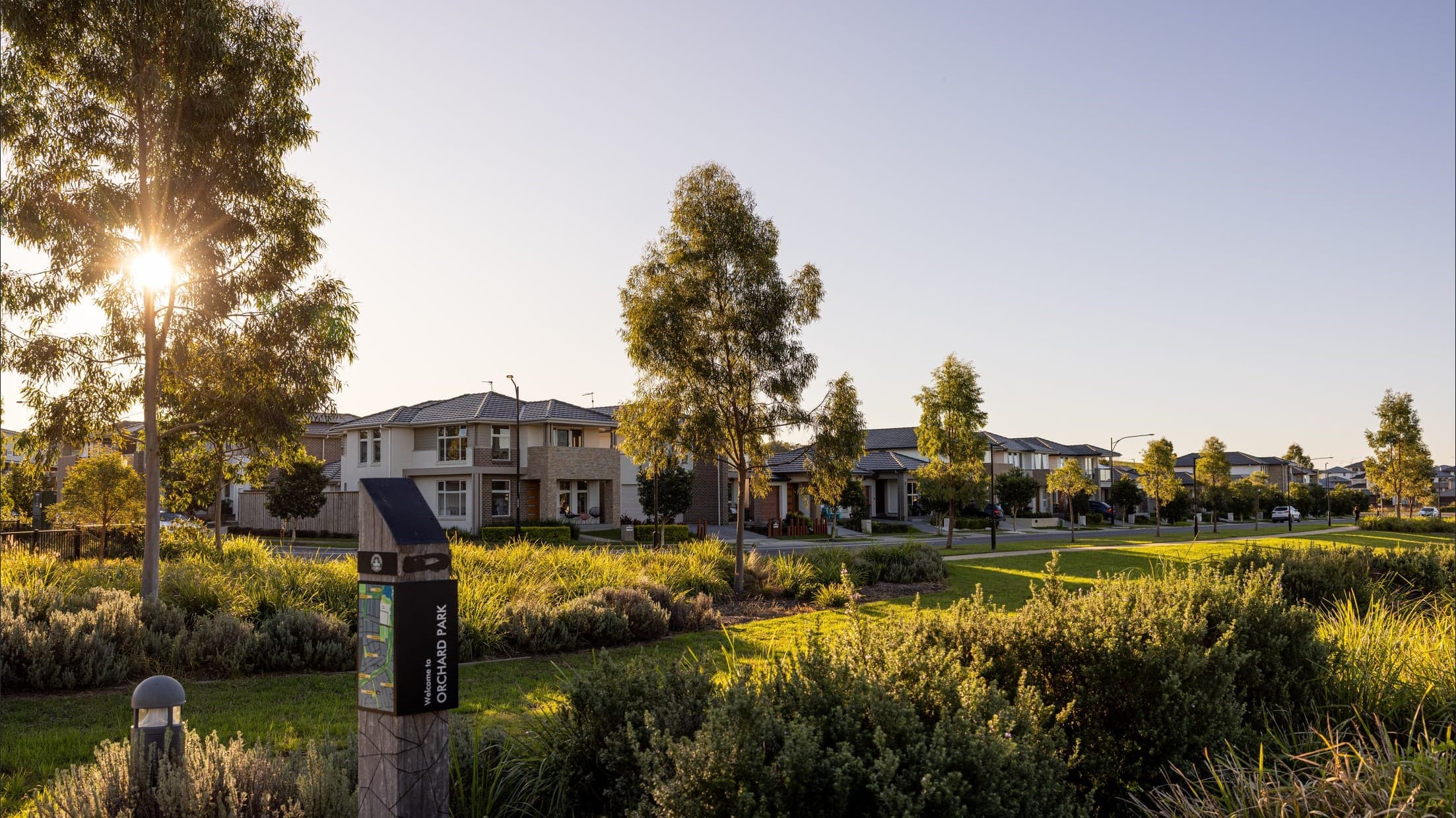 A suburban neighborhood at sunset featuring modern houses, neatly trimmed lawns, trees, and a park sign labeled Orchid Park surrounded by lush greenery. The sun is partially visible behind the trees.