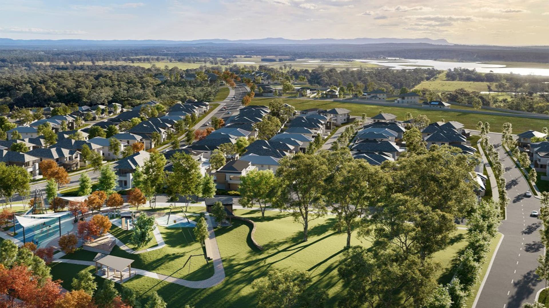 Aerial view of a suburban neighborhood with houses, tree-lined streets, green parks, walking paths, and a river in the background under a partly cloudy sky.