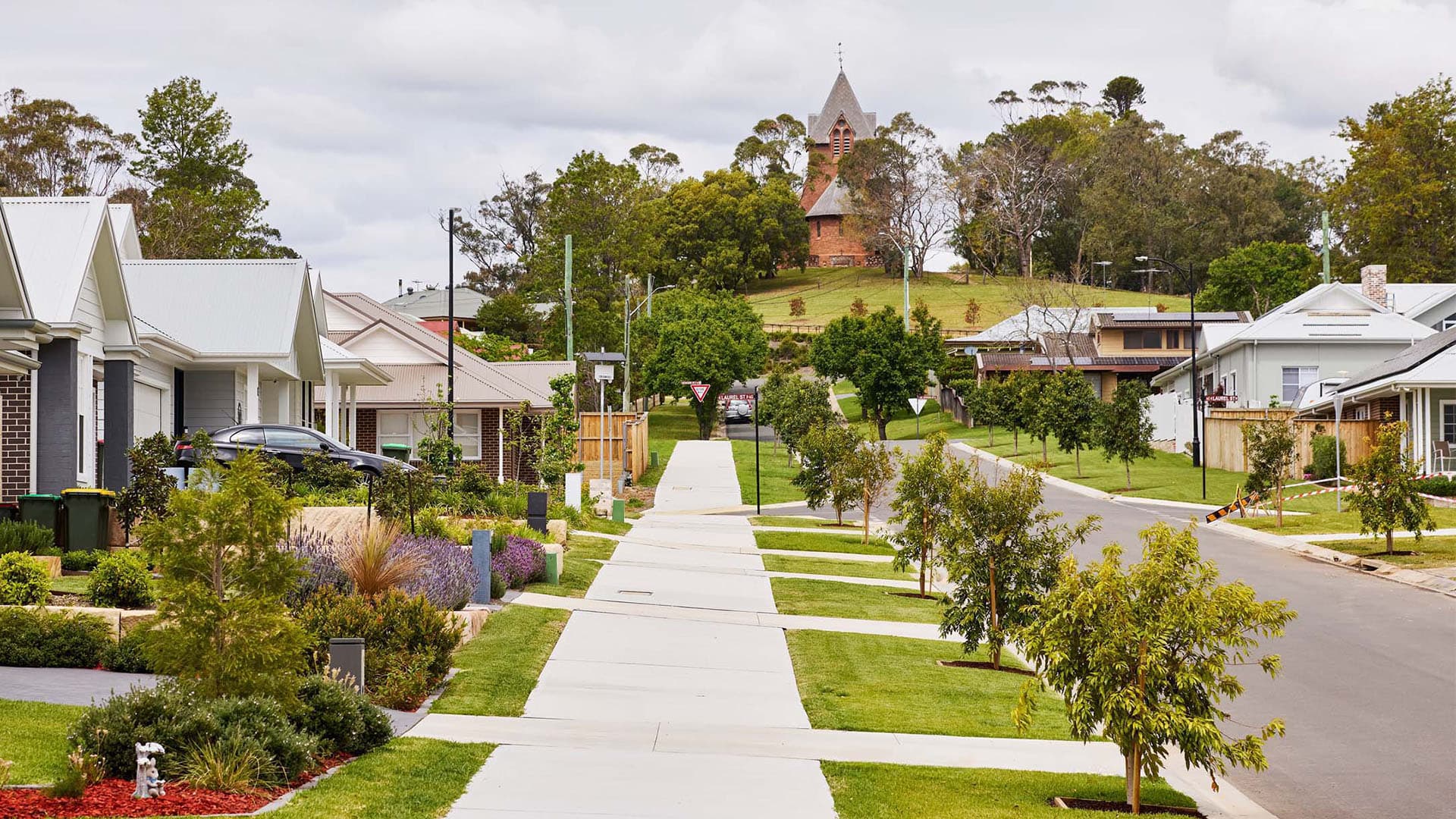 A quiet suburban street with modern houses, well-kept lawns, and young trees lining the sidewalk. In the background, a historic church with a pointed steeple sits atop a grassy hill surrounded by trees.