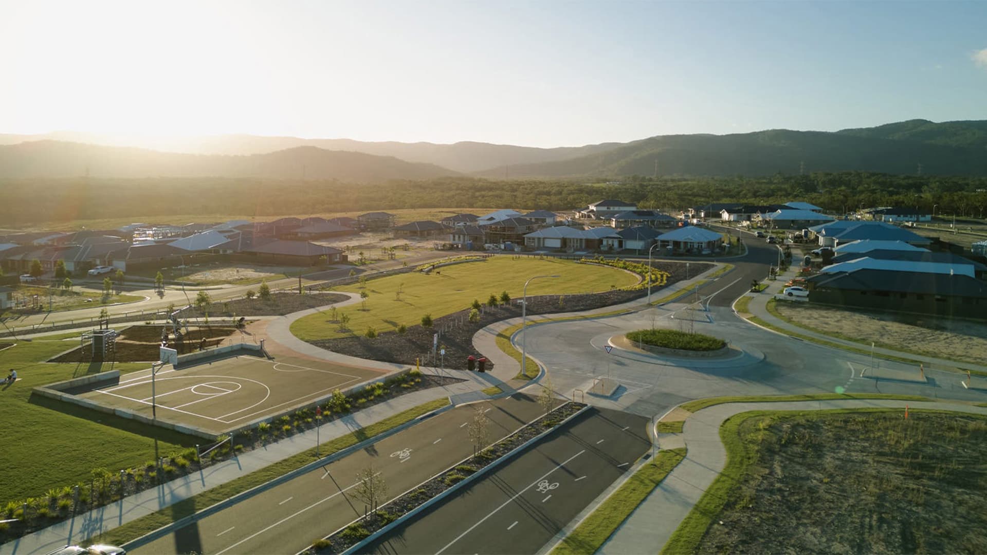 Aerial view of a suburban neighborhood at sunset, featuring curving streets, houses with blue roofs, a basketball court, a playground, and green open spaces; mountains are visible in the background.