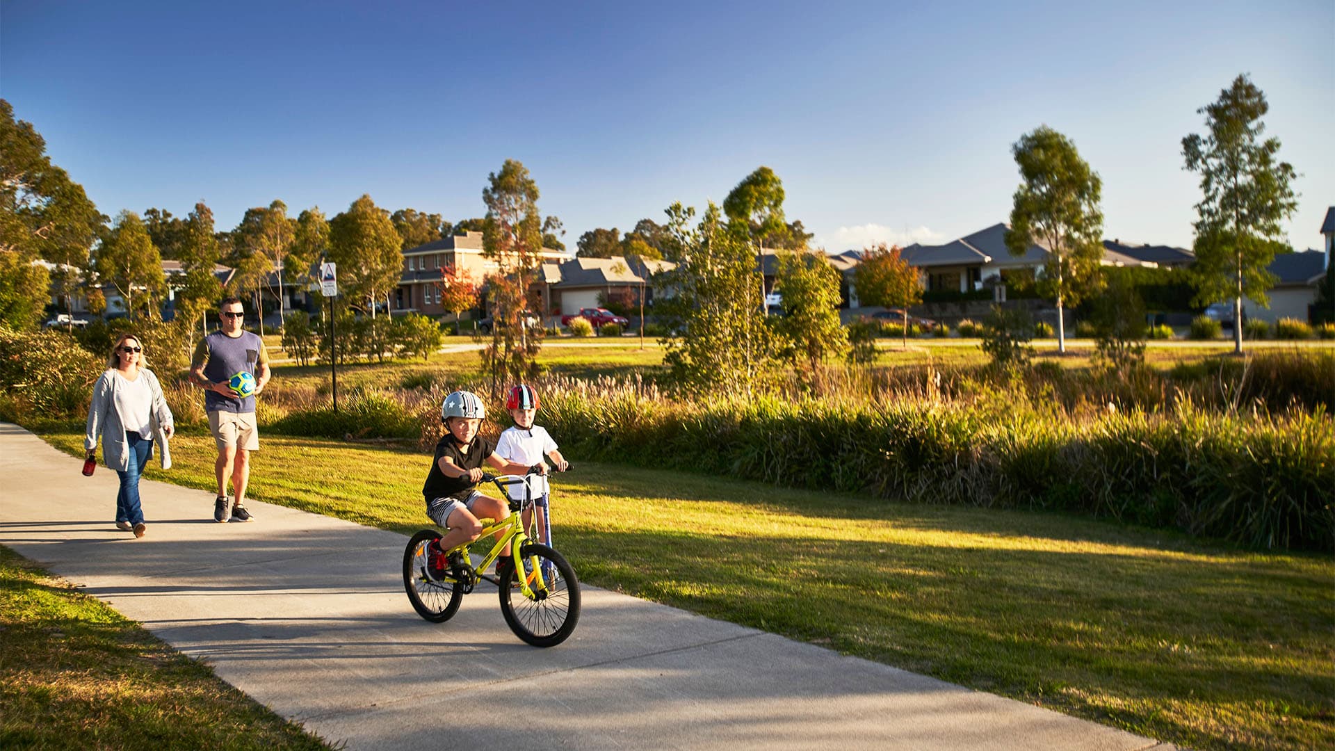 A young child rides a bike on a sunny path while another child walks beside them. Two adults walk behind, enjoying the day in a suburban neighborhood with houses and trees in the background.