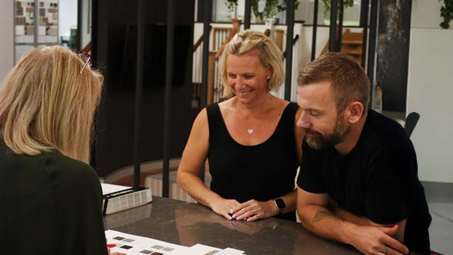 Two people sit at a table, smiling and looking at papers or color samples while another person, with back to camera, discusses options with them in a modern office setting.