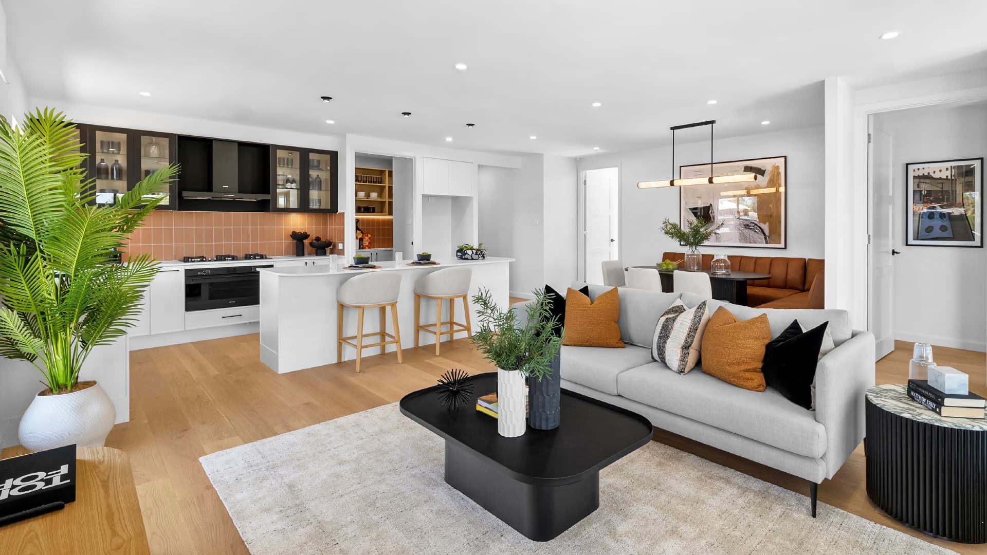 Modern open-plan living space with a kitchen, dining area, and living room. Features a white island, black accents, a gray sofa with orange cushions, wooden floors, plants, and contemporary decor.