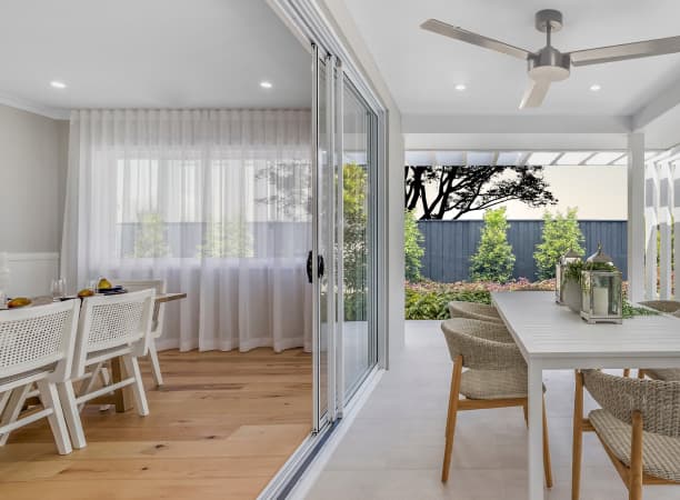 A modern dining area with white furniture flows into a covered patio with outdoor seating. Large sliding glass doors separate the indoor space from the patio, and greenery is visible outside the black fence.