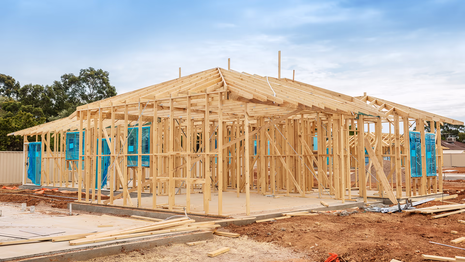A single-story house under construction, featuring a wooden frame structure with exposed beams and blue plastic coverings on window openings, set on a dirt lot under a partly cloudy sky.