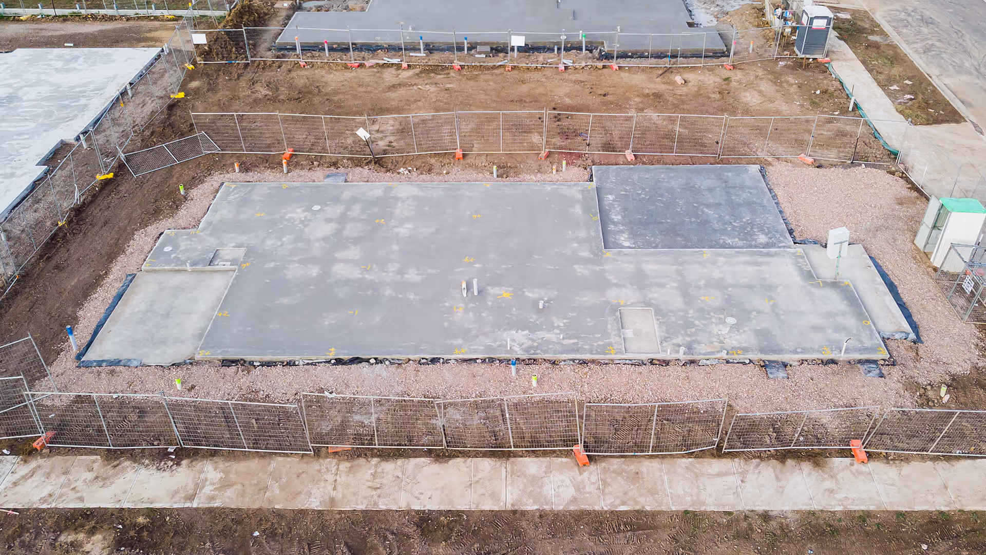 Aerial view of a building foundation under construction, surrounded by temporary fencing, gravel, and bare soil, with concrete slabs and construction materials visible on the site.