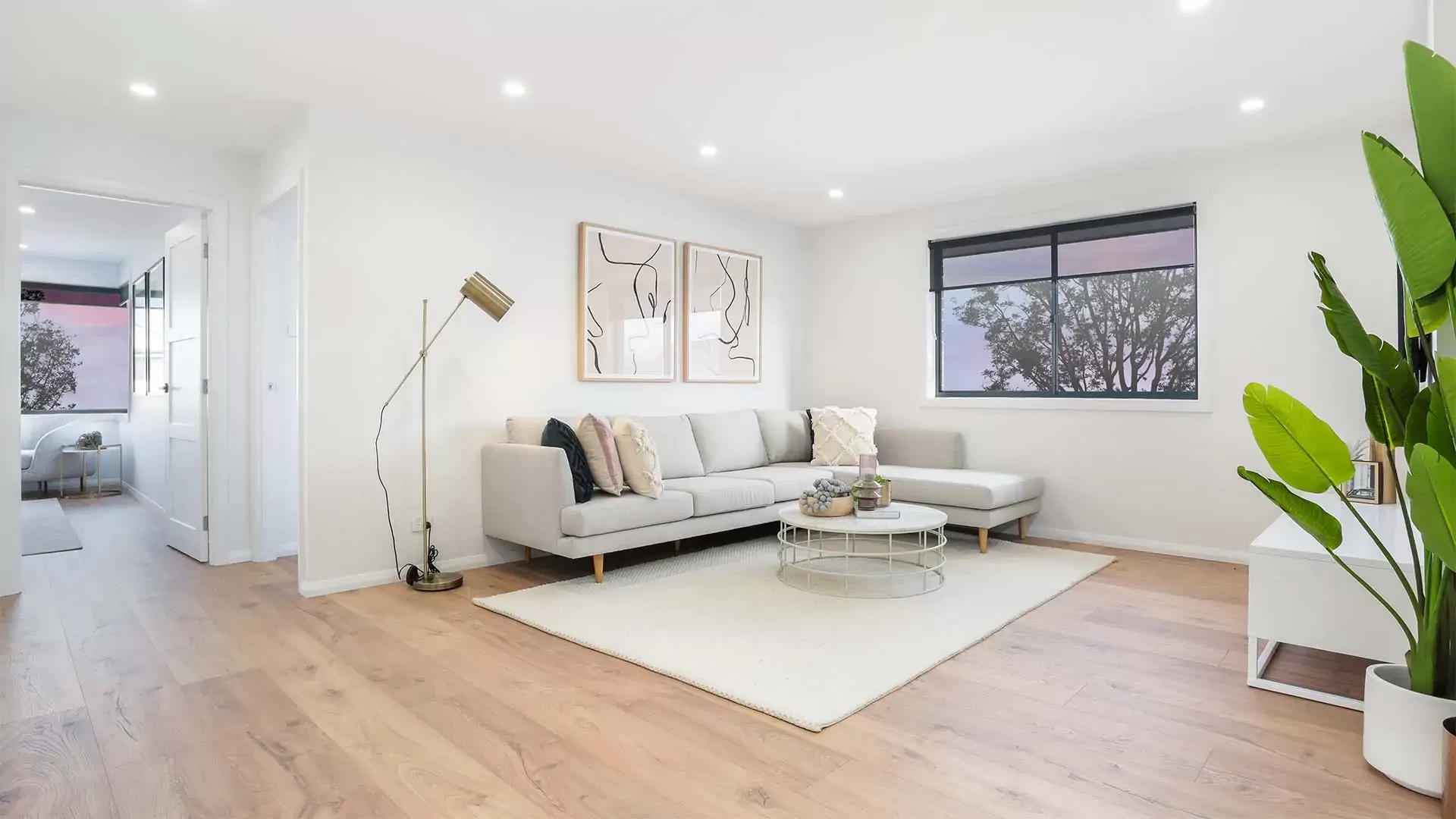 Modern living room with light wood floors, a gray sectional sofa, abstract wall art, a floor lamp, round coffee tables, large indoor plant, and a window with a view of trees. Natural light fills the bright, minimal space.