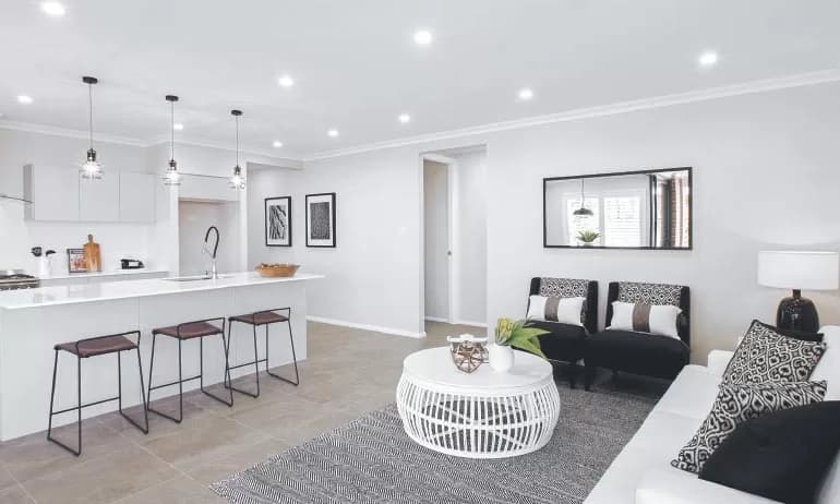 Modern open-plan kitchen and living room with white walls, pendant lights, a breakfast bar with stools, black and white furniture, decorative cushions, a round coffee table, and a patterned rug.