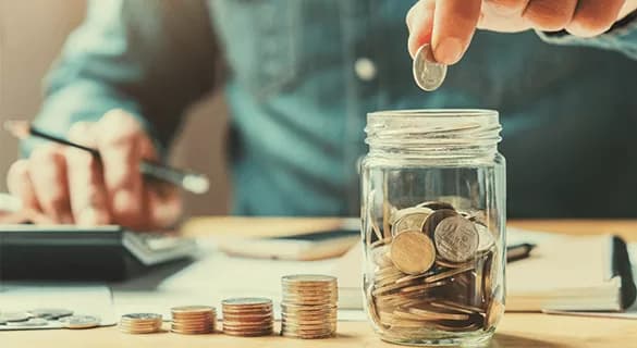 A person drops a coin into a glass jar filled with coins. Stacks of coins, papers, and a calculator are on the table, suggesting budgeting or saving money. The persons face is not visible.