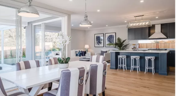 Modern open-plan kitchen and dining area with a white dining table, striped chairs, potted orchid centerpiece, kitchen island with barstools, pendant lights, wood flooring, and large windows providing natural light.