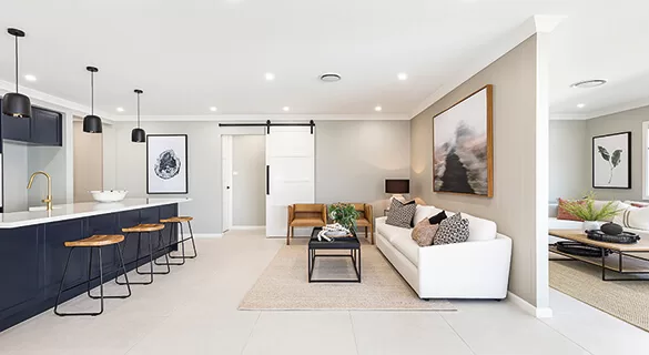 Modern open-plan living space with a kitchen featuring navy cabinets and barstools, a white sofa, minimalist decor, large abstract art, and neutral tones throughout. A sliding barn door separates rooms.