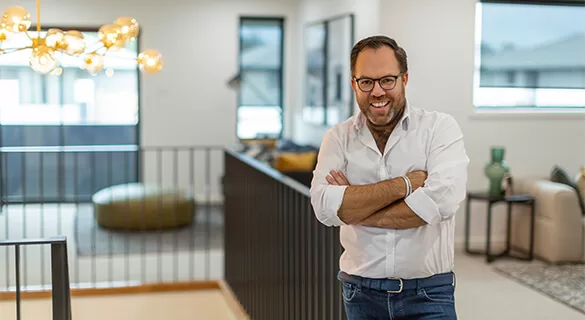 A man with glasses, wearing a white shirt and blue jeans, stands indoors with his arms crossed, smiling. The modern room has large windows, a couch, a table, and hanging lights in the background.