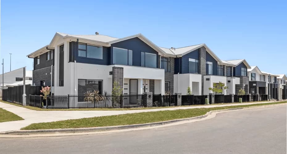 Modern two-story townhouses with large windows and balconies, lined up along a clean, quiet street with a grassy verge and sidewalk under a clear blue sky.