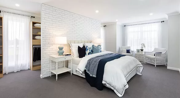Spacious, modern bedroom with a white brick accent wall, large bed with white and navy bedding, two bedside tables with lamps, open closet, and two white armchairs near large windows with sheer curtains.
