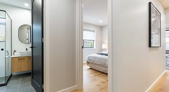 A hallway with light-colored walls leads to a modern bathroom on the left, featuring a glass shower and wood vanity, and a bright bedroom with a bed, lamp, and large window straight ahead.