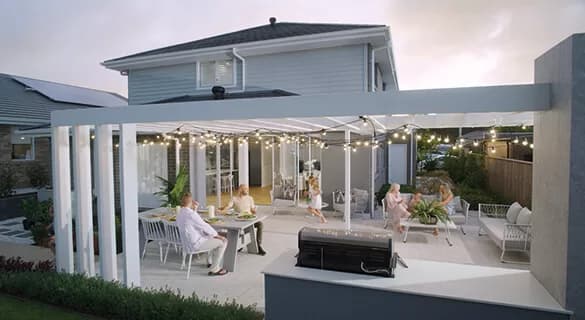 A modern outdoor patio with pergola and string lights, attached to a two-story house. Several people are sitting and dining at tables, enjoying a meal and conversation in a relaxed setting.