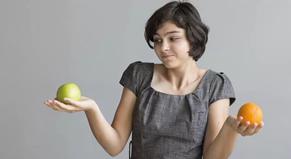 A woman in a gray dress holds a green apple in one hand and an orange in the other, looking at them with a thoughtful expression against a plain gray background.