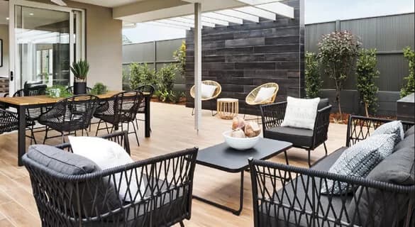 Modern outdoor patio with black wicker chairs, gray cushions, a black coffee table, potted plants, a wooden dining table, and decorative pillows, set on a wooden deck with privacy fencing and greenery around.