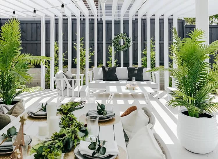 A modern outdoor patio with white pergola, lush green plants, a dining table set with dishes and greenery, and a white sofa with dark cushions under dappled sunlight. A green wreath hangs on the pergola slats.