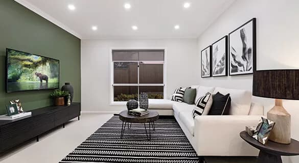 Modern living room with white walls, one green accent wall, a white sectional sofa with black and white pillows, a striped rug, a TV, coffee table, table lamp, and framed artwork above the sofa. Ceiling lights are on.