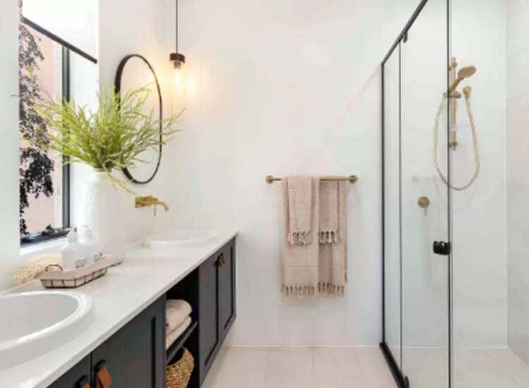 Modern bathroom with a glass shower, double sinks, large mirror, black cabinets, gold fixtures, a hanging light, beige towels on a towel bar, and a vase with green foliage on the counter.
