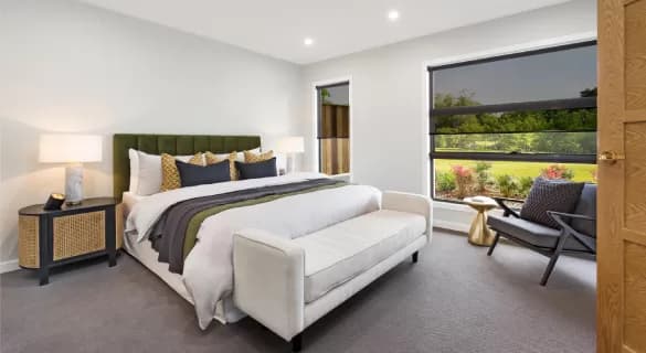 Modern bedroom with a large bed, white bench, two nightstands with lamps, a black armchair, and large window overlooking a green outdoor area. Room features neutral colors and contemporary decor.